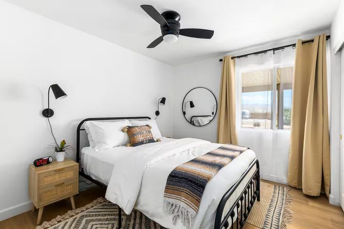 Bright modern bedroom with black metal bedframe, white bedding, striped woven throw, round wall mirror, wood nightstand, black wall lamps, tan curtains and ceiling fan