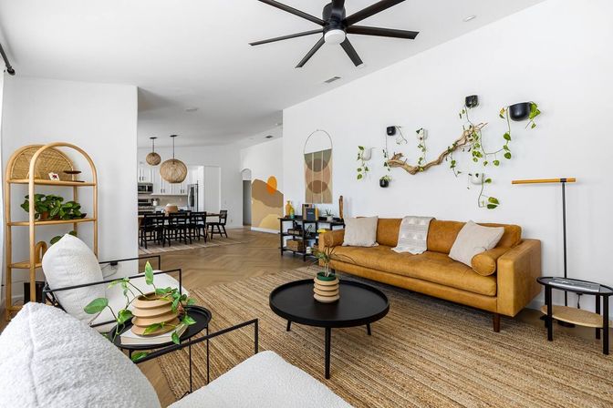 Bright open-concept living room with tan leather sofa, woven jute rug, black round coffee table, hanging vine on driftwood wall display, rattan shelving, herringbone hardwood floors and dining area with pendant lights in background.