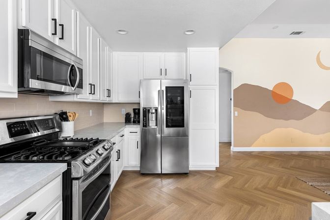 Open-plan modern kitchen interior with white shaker cabinets, stainless steel gas range, microwave and French-door refrigerator, light subway tile backsplash, herringbone wood floor and warm sunset mural on the adjacent wall.