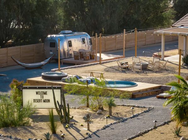 Sunlit desert glamping courtyard with a silver Airstream trailer, hammock, wooden deck with dining area, round plunge pool and hot tub, fire-pit seating under string lights, gravel paths and cacti with a welcome sign in the foreground.