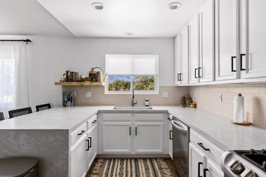 Bright modern white kitchen with U-shaped quartz countertops, farmhouse sink under window with tree view, black cabinet hardware, stainless dishwasher and patterned runner