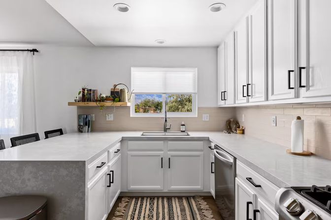 Bright modern white kitchen with U-shaped quartz countertops, farmhouse sink under window with tree view, black cabinet hardware, stainless dishwasher and patterned runner