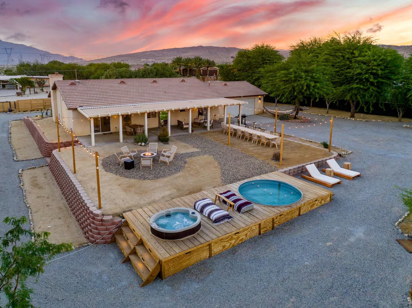 Cozy desert backyard retreat at sunset: single-story house with string lights, long outdoor dining table, circular fire pit, wooden deck with hot tub and plunge pool, lounge chairs and mountain views.