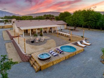 Cozy desert backyard retreat at sunset: single-story house with string lights, long outdoor dining table, circular fire pit, wooden deck with hot tub and plunge pool, lounge chairs and mountain views.