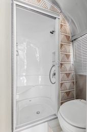 Compact white shower stall with sliding door, built-in corner seat and handheld chrome showerhead, brown geometric tile accent, window blinds and adjacent toilet.