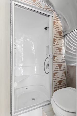 Compact white shower stall with sliding door, built-in corner seat and handheld chrome showerhead, brown geometric tile accent, window blinds and adjacent toilet.