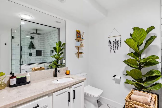 Bright modern bathroom interior with white subway-tiled walk-in shower, marble countertop vanity, matte black fixtures, large mirror and fiddle-leaf fig plant
