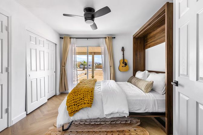 Bright modern bedroom with wood-framed bed alcove, white linens and mustard knit throw, wall-mounted acoustic guitar, ceiling fan, and sliding glass door opening to a sunny patio with distant mountain view.