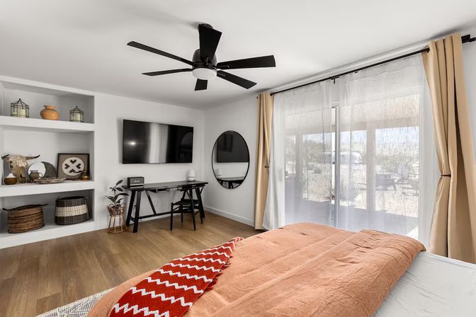 Cozy sunlit modern bedroom with rust-orange duvet and red chevron throw, wall-mounted TV, built-in shelves with baskets and decor, black ceiling fan, desk with round mirror, and sheer-curtained sliding glass doors opening to a patio/desert view.