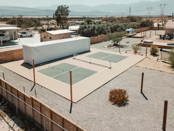 Aerial view of two sun‑baked pickleball courts in a fenced desert backyard with string lights, a white shipping container, a silver camper, gravel landscaping and distant mountains under a clear sky.