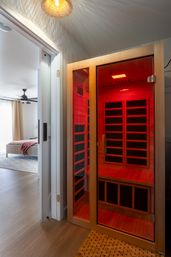 Cozy modern bedroom with an adjoining wooden infrared sauna glowing red through a glass door — home spa bench, hardwood floors, and ambient lighting.