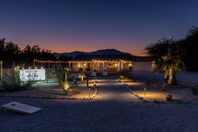 Cozy desert yard at twilight with illuminated gravel path, string lights draped across a single-story home, palm trees and mountain silhouettes in the background.