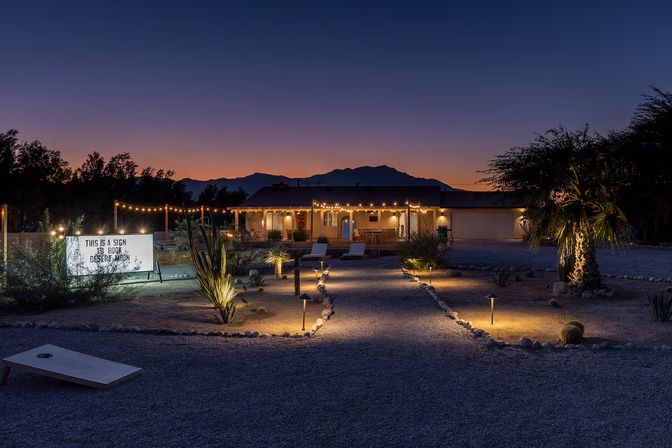 Cozy desert yard at twilight with illuminated gravel path, string lights draped across a single-story home, palm trees and mountain silhouettes in the background.