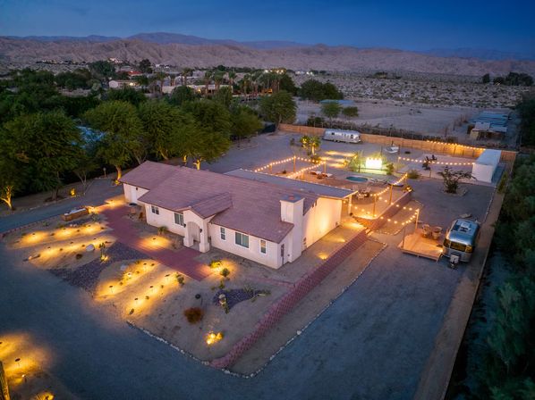 Aerial dusk view of a desert oasis: lit stucco home with string lights, patio and hot tub, Airstream trailers, gravel landscaping and mountains beyond.