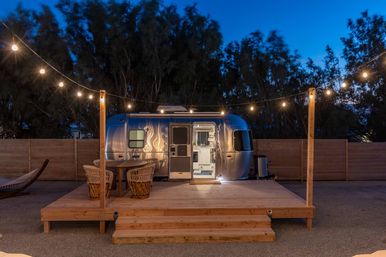 Silver retro travel trailer on a raised wooden deck at dusk, cozy outdoor glamping setup with hanging string lights, wicker dining chairs and table, and a nearby hammock