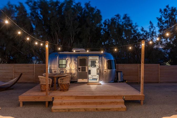 Silver retro travel trailer on a raised wooden deck at dusk, cozy outdoor glamping setup with hanging string lights, wicker dining chairs and table, and a nearby hammock