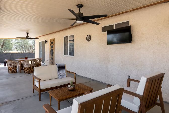 Covered backyard patio with wood-framed cushioned seating, wooden coffee table with a small cactus, ceiling fans, wall-mounted TV and a dining table with woven chairs overlooking trees