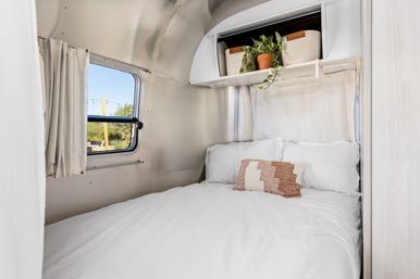 Cozy white bed in an aluminum-paneled travel trailer bedroom with a textured throw pillow, potted plant on an overhead shelf, and a small window looking out to a sunny RV park view.