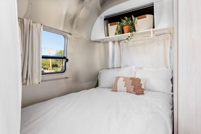 Cozy white bed in an aluminum-paneled travel trailer bedroom with a textured throw pillow, potted plant on an overhead shelf, and a small window looking out to a sunny RV park view.