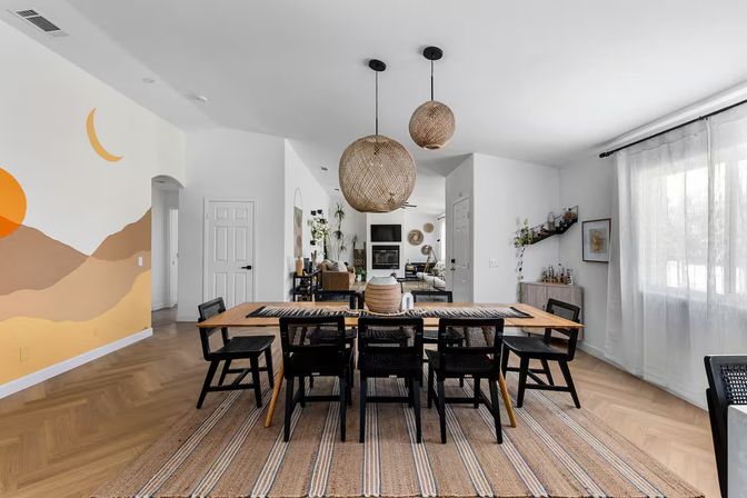 Sunlit open-concept boho-modern dining room with a long wooden table and six black woven chairs, oversized rattan pendant lights, striped jute rug on herringbone hardwood floor, warm desert mural with sun and crescent moon on left wall, sheer curtains and a cozy living area with fireplace and plants in the background.