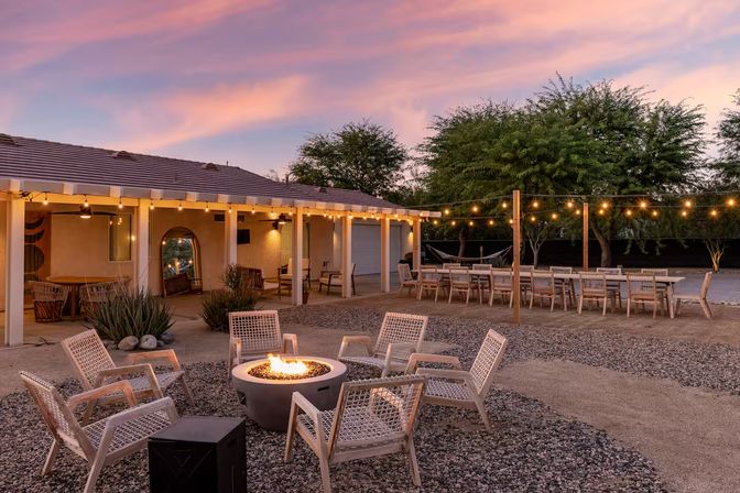 Sunset desert backyard patio with a glowing fire pit surrounded by woven lounge chairs, long outdoor dining table under string lights, covered porch and trees.