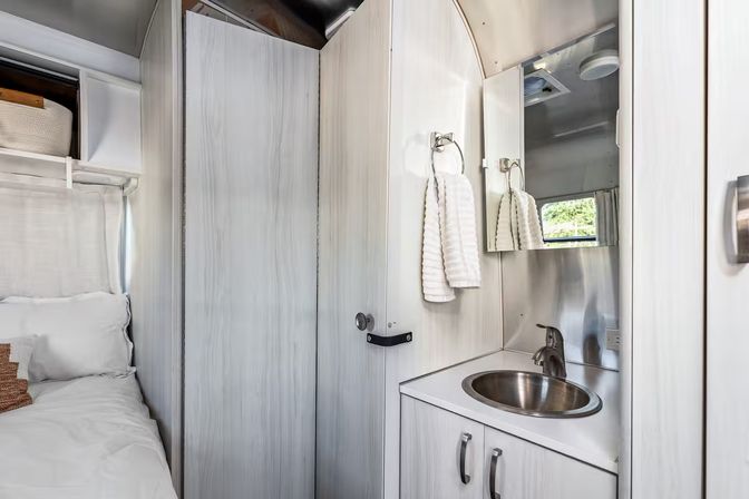 Cozy, bright RV bathroom with stainless-steel sink and faucet, mirrored medicine cabinet, white hand towel on a ring, and a bed visible beside light wood cabinetry.