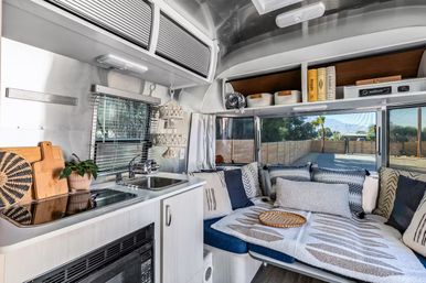 Cozy modern travel trailer interior featuring a compact stainless-steel kitchenette with sink and stovetop, microwave and overhead storage, plus a cushioned lounge/bed with patterned pillows and throws beside large windows overlooking a sunny palm-tree yard.