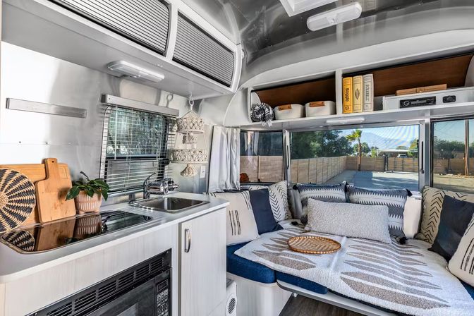 Cozy modern travel trailer interior featuring a compact stainless-steel kitchenette with sink and stovetop, microwave and overhead storage, plus a cushioned lounge/bed with patterned pillows and throws beside large windows overlooking a sunny palm-tree yard.