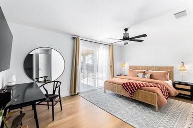 Sunlit modern bedroom with king-size wicker platform bed in rust bedding and patterned throw, large geometric area rug on wood floors, black ceiling fan, sliding glass doors with sheer curtains, round wall mirror and black desk workspace.