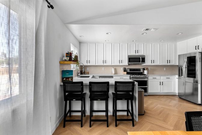 Sunlit open-concept modern white kitchen with a large island and three black bar stools, stainless steel fridge and oven, white shaker cabinets, light subway-tile backsplash, and herringbone wood floor by a sheer-curtained window.