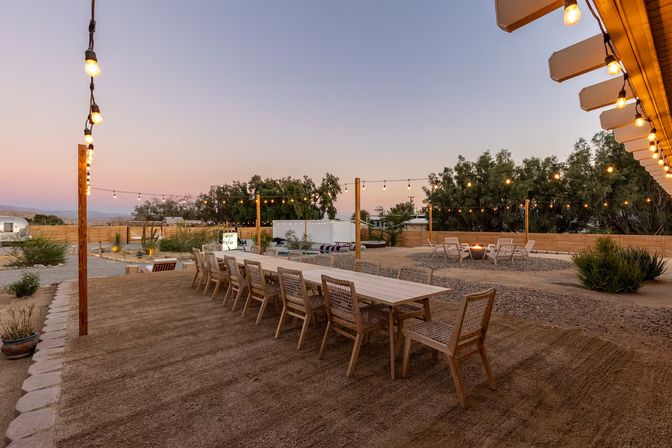 Cozy outdoor desert patio at sunset featuring a long wooden communal table with wicker chairs beneath warm string lights, gravel courtyard with a circular fire-pit seating area and distant hills