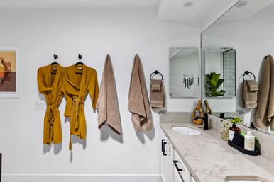 Modern white bathroom with marble countertop and large mirror; black fixtures, two cheerful mustard waffle-knit bathrobes and beige towels on wall hooks, potted plant and toiletries on the vanity.