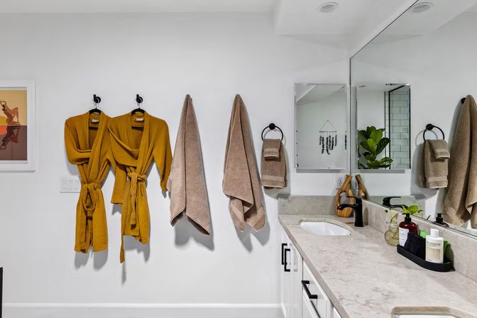 Modern white bathroom with marble countertop and large mirror; black fixtures, two cheerful mustard waffle-knit bathrobes and beige towels on wall hooks, potted plant and toiletries on the vanity.