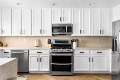 Bright modern white kitchen with shaker cabinets, stainless-steel gas range and over-the-range microwave, dishwasher and fridge, beige subway tile backsplash and herringbone wood floor.