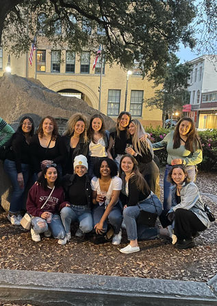 Group of 13 friends posing outdoors at dusk in a downtown plaza — smiling women in casual outfits gathered around a large rock monument in front of a historic brick building under an oak tree.
