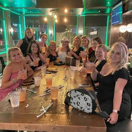 Smiling group of women raising cocktails around a long wooden table in a neon-lit bar, one wearing a white cowboy hat — girls' night celebration with plastic cups and handbags on the table.