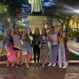 Seven friends smiling and toasting with drinks in front of a historic statue on a tree-lined downtown plaza at dusk, lively nighttime city scene
