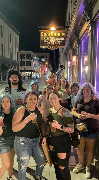 Group of friends in colorful wigs and casual clothes smiling and holding drinks on a downtown sidewalk at night outside a neon-lit bar, lively nightlife street scene.