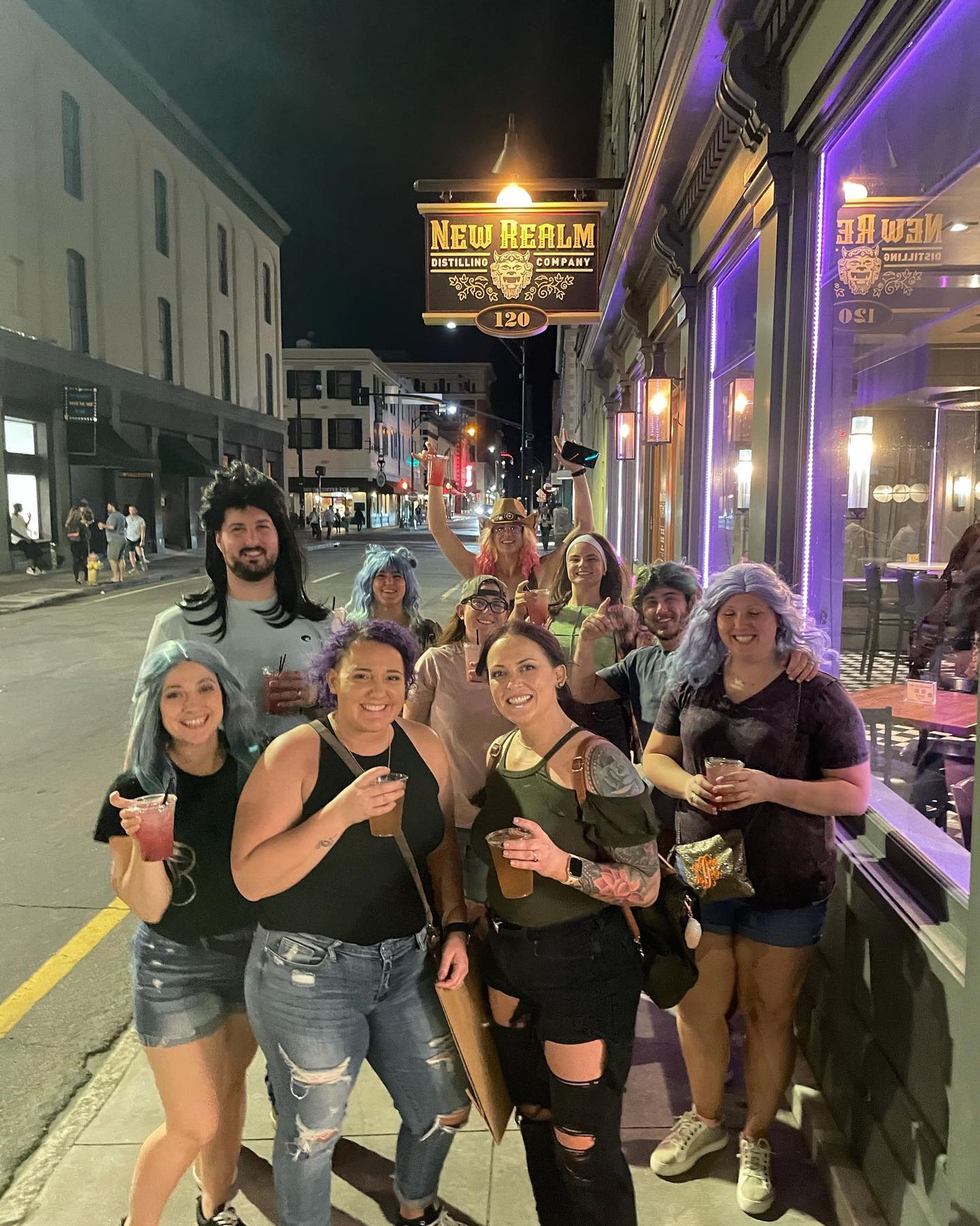 Group of friends in colorful wigs and casual clothes smiling and holding drinks on a downtown sidewalk at night outside a neon-lit bar, lively nightlife street scene.