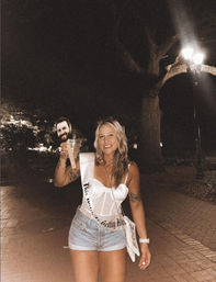Smiling woman in a white corset top, denim shorts and bachelorette sash holding a cocktail on a nighttime brick walkway under a streetlamp, companion smiling in the background — nightlife bachelorette celebration
