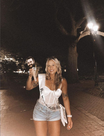Smiling woman in a white corset top, denim shorts and bachelorette sash holding a cocktail on a nighttime brick walkway under a streetlamp, companion smiling in the background — nightlife bachelorette celebration