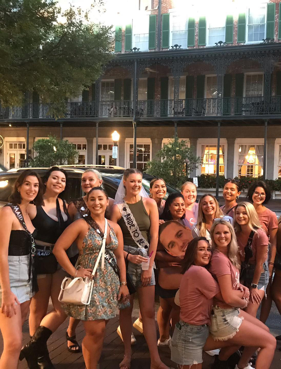 Smiling bachelorette party of women on a historic downtown street with wrought-iron balconies, bride-to-be in veil and sash.