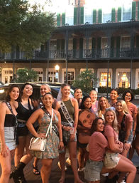 Smiling bachelorette party of women on a historic downtown street with wrought-iron balconies, bride-to-be in veil and sash.