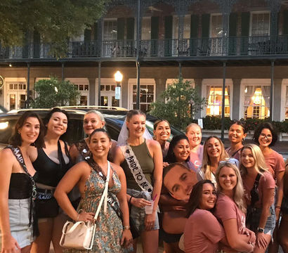 Smiling bachelorette party of women on a historic downtown street with wrought-iron balconies, bride-to-be in veil and sash.