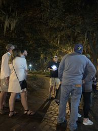 Night walking tour: a small group gathered on a brick pathway beneath live oak trees draped in Spanish moss as a guide holds an illuminated tablet to point out sights.