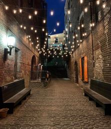 Twilight cobblestone alley flanked by brick buildings and benches, overhead string lights creating a cozy urban ambiance with stairs at the far end.