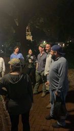 Group of friends sharing drinks and laughs on a brick sidewalk under trees at night in a downtown park.