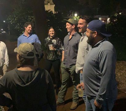 Group of friends sharing drinks and laughs on a brick sidewalk under trees at night in a downtown park.