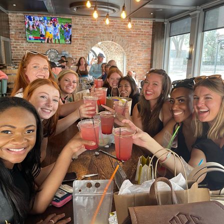 Group of smiling women toasting pink cocktails around a long wooden table in a brick-walled bar with hanging Edison bulbs and large windows — lively daytime gathering of friends.
