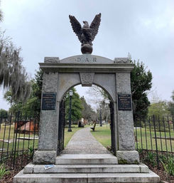 Granite archway entrance topped by a soaring eagle statue leading into a historic cemetery — iron fence, lamp posts, moss-draped oaks and a winding gravel path.